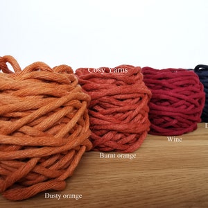 K&ouml;nnte beinhalten: Vier Garnrollen in verschiedenen Farben: staubiges Orange, gebranntes Orange, Wein und Dunkelblau. Das Garn ist in einer Reihe auf einer Holzoberfl&auml;che angeordnet. Der Text "Cosy Yarns" ist auf dem Etikett der gebrannten orangen Garnrolle sichtbar.