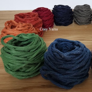 May include: Close-up of eight balls of yarn in various colours, including orange, red, blue, grey, and green. The yarn is arranged in a circle on a wooden surface. The text "Cosy Yarns" is visible in the centre of the image.
