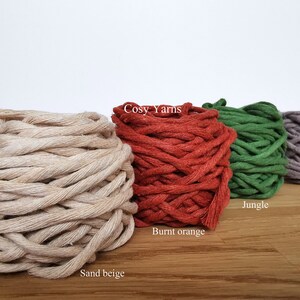 K&ouml;nnte beinhalten: F&uuml;nf Garnrollen in verschiedenen Farben: Sandbeige, gebranntes Orange, Dschungelgr&uuml;n, Mokka und ein wei&szlig;es Etikett mit der Aufschrift "Cosy Yarns".