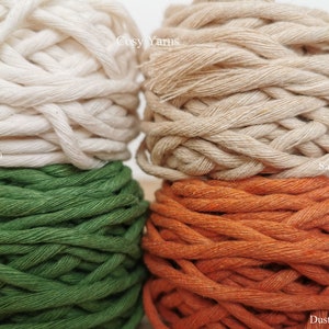 May include: Four spools of cotton rope in different colors: light ecru, jungle green, dusty orange, and sand beige. The spools are stacked on top of each other. The text "Cosy Yarns" is visible on the top spool.