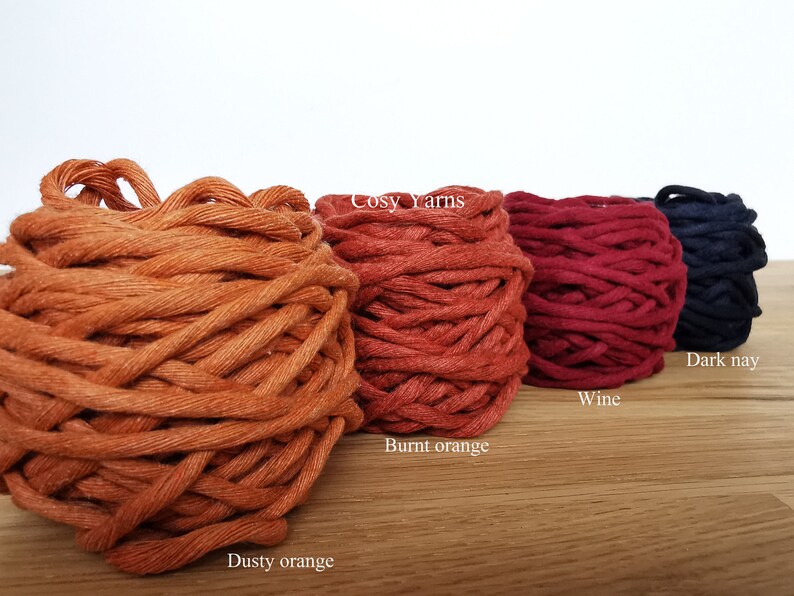 May include: Four spools of yarn in different colors: dusty orange, burnt orange, wine, and dark navy. The yarn is arranged in a row on a wooden surface. The text "Cosy Yarns" is visible on the label of the burnt orange spool.