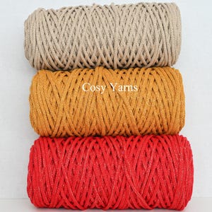 May include: Three spools of yarn in beige, gold, and red. The gold spool has the text "Cosy Yarns" printed on it. The yarn is tightly wound, showing a textured surface. The red yarn has a metallic sheen.