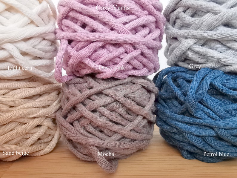 K&ouml;nnte beinhalten: Sechs Spulen mit Baumwollgarn in verschiedenen Farben, darunter helles Ecru, Sandbeige, staubiges Rosa, Mokka, Grau und Petrolblau. Die Spulen sind in zwei Reihen zu je drei angeordnet, wobei der Text "Cosy Yarns" &uuml;ber der oberen Reihe gedruckt ist.