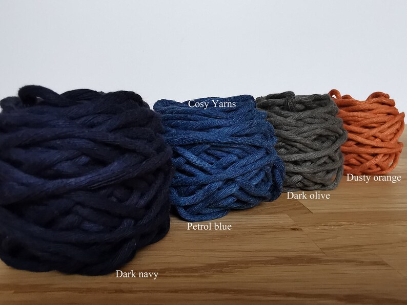 K&ouml;nnte beinhalten: Vier Kn&auml;uel Garn in verschiedenen Farben: Dunkelblau, Petrolblau, Dunkeloliv und Stauborange. Das Garn ist mit "Cosy Yarns" gekennzeichnet.