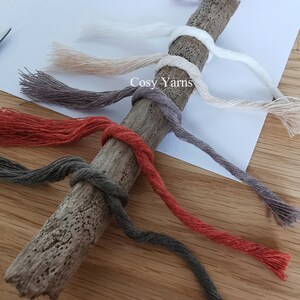 K&ouml;nnte beinhalten: Eine Sammlung bunter Garnstr&auml;nge, die um ein St&uuml;ck Treibholz gewickelt sind. Die Garnfarben sind rot, orange, braun, grau, beige und wei&szlig;. Der Text "Cosy Yarns" ist auf dem Treibholz sichtbar.