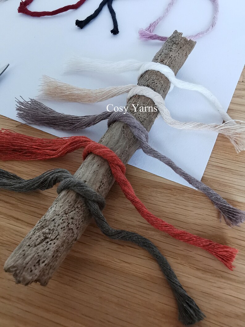 K&ouml;nnte beinhalten: Eine Sammlung bunter Garnstr&auml;nge, die um ein St&uuml;ck Treibholz gewickelt sind. Die Garnfarben sind rot, orange, braun, grau, beige und wei&szlig;. Der Text "Cosy Yarns" ist auf dem Treibholz sichtbar.