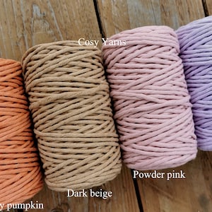 May include: Four spools of macrame cord in shades of dusty pumpkin, dark beige, powder pink, and lilac. The text "Cosy Yarns" is visible. The tightly wound spools are displayed on a wooden surface.