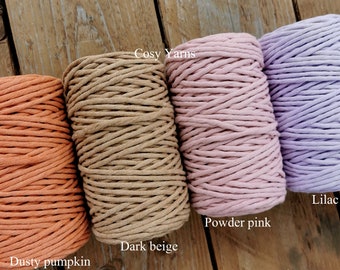May include: Four spools of macrame cord in various colors: dusty pumpkin, dark beige, powder pink, and lilac. The text "Cosy Yarns" is visible at the top. The spools are tightly wound and sit on a wooden surface.