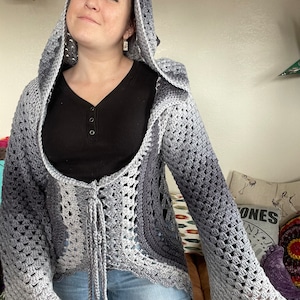 Puede incluir: Un cárdigan de crochet gris y blanco degradado con capucha y mangas acampanadas. El cárdigan está atado a la cintura con un cordón largo.