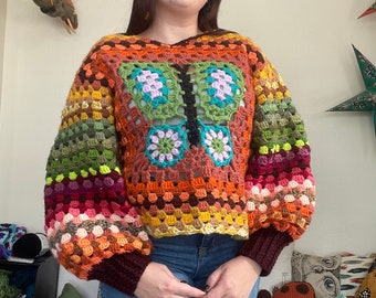 Könnte beinhalten: Ein bunter Häkelpullover mit einem Schmetterlingsmuster. Der Pullover ist aus verschiedenen Farben gefertigt, darunter Rot, Orange, Gelb, Grün, Blau und Lila. Der Pullover hat eine lockere Passform und lange Ärmel.