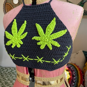 Może przedstawiać: Czarny top halterowy wykonany na szydełku z zielonymi wzorami liści marihuany. Top ma złoty pasek wokół dekoltu.