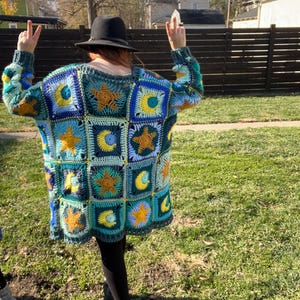 May include: A handmade, oversized crochet sweater in shades of teal, blue, yellow, and gold. The sweater features a patchwork design with squares adorned with stars and crescent moons. The sleeves are long and the overall style is relaxed.