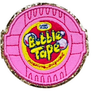 May include: A pink embroidered patch with the words "Bubble Tape" in yellow and red, surrounded by a gold glitter border. The patch has a retro design, reminiscent of vintage candy packaging.