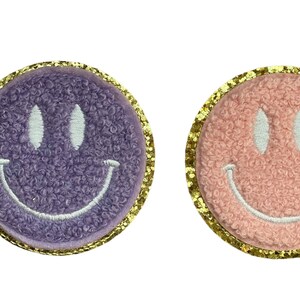 May include: Two fuzzy smiley face patches with gold glitter trim. One patch is purple and the other is pink.