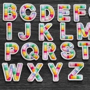 May include: A full alphabet of embroidered patches, each letter in a different design. The letters are in a rainbow of colors, with a white border. The patches are on a dark gray wooden surface.