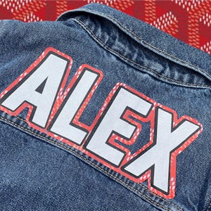 Könnte beinhalten: Jeansjacke mit dem Namen "ALEX" in großen weißen Buchstaben, umrandet von Rot und Schwarz. Die Buchstaben haben ein Muster. Die Jacke befindet sich auf einem rot-weiß gemusterten Hintergrund.