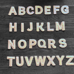 May include: A set of 26 iridescent alphabet letters, each letter is individually cut out and has a white outline. The letters are arranged in alphabetical order in four rows on a dark wood surface.
