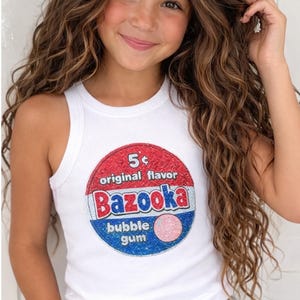 May include: White ribbed tank top with a large, colorful "Bazooka bubble gum" graphic. The graphic features red, white, and blue elements with the text "5¢ original flavor". The tank top is worn by a person.
