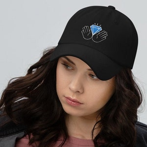 May include: Black baseball cap with a diamond and hands embroidered design. The cap features a curved brim and an adjustable strap for a comfortable fit. The diamond is blue with white highlights, and the hands are outlined in blue.