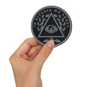 May include: A circular black patch with a grey border, featuring the text "IN BITCOIN WE TRUST" around a triangle with an eye symbol. The patch is held in a hand against a white backdrop.