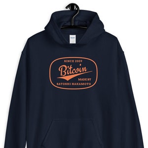 Puede incluir: Una sudadera con capucha azul marino con un logotipo naranja de estilo vintage que dice "Bitcoin" con el texto "Since 2009" y "Made by Satoshi Nakamoto" debajo.