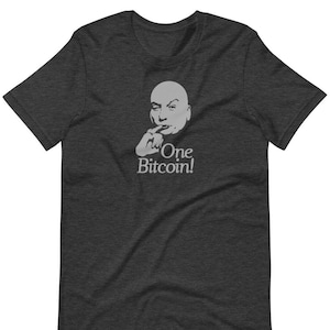 Puede incluir: Una camiseta gris oscuro jaspeada con una imagen blanca de un hombre calvo con una expresión seria. El hombre lleva una camisa blanca y tiene el dedo en los labios. El texto "One Bitcoin!" está impreso debajo de la imagen.