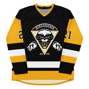 Könnte beinhalten: Ein schwarzes und goldenes Hockeytrikot mit einem weißen Dreieckslogo, das einen Honigdachs mit dem Text "Honey Badger" und "2009" zeigt. Das Trikot hat weiße und schwarze Streifen an den Ärmeln und einen weißen Streifen am unteren Rand.