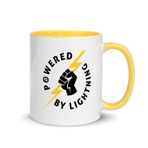 Puede incluir: Taza de cerámica blanca con borde y asa amarillos. La taza presenta un puño negro sujetando un rayo amarillo con el texto "Powered by Lightning" en un círculo alrededor de la imagen.