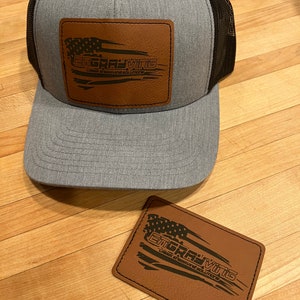 May include: A gray and black trucker hat with a brown leather patch that says "Engraving - Laser Engraved Solutions". The patch features a stylized American flag.