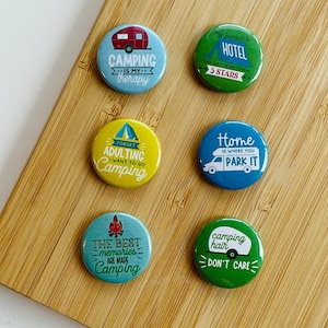 Könnte beinhalten: Sechs bunte runde Knöpfe mit Sprüchen zum Thema Camping. Die Knöpfe befinden sich auf einer Holzoberfläche. Die Sprüche auf den Knöpfen lauten: "Camping ist meine Therapie", "Mein Lieblingshotel hat weit mehr als 5 Sterne", "Vergiss das Erwachsenwerden, ich will campen gehen", "Zuhause ist, wo man parkt", "Die besten Erinnerungen entstehen beim Campen", und "Camping-Haare, egal".