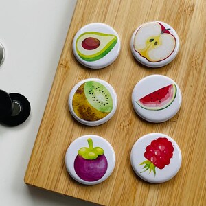 Könnte beinhalten: Sechs runde weiße Magnete mit bunten Bildern von Früchten. Die Magnete zeigen einen Kiwi, eine Avocado, eine Wassermelone, einen Apfel, eine Mangostane und Himbeeren.