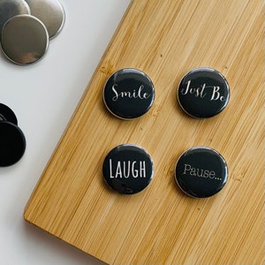 Könnte beinhalten: Eine Sammlung schwarzer runder Knöpfe mit weißem Text, darunter "Smile", "Just Be", "Laugh" und "Pause...". Zusätzliche silberne und schwarze Knöpfe liegen auf einer Holzoberfläche. Die Knöpfe dienen wahrscheinlich dekorativen oder funktionalen Zwecken.