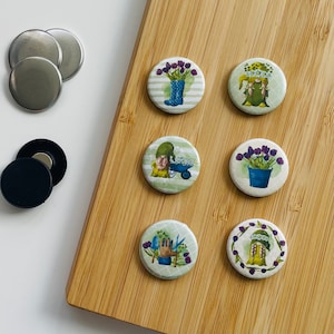 Könnte beinhalten: Satz von sieben runden Magneten mit Gartenthema. Die Magnete zeigen Bilder von Gnom, Blumen, Gartengeräten und einem Gnom mit einer Blumenkrone. Die Magnete befinden sich auf einer Holzoberfläche.