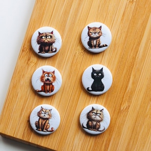 Peut inclure: Six boutons ronds blancs avec des illustrations de chats de dessin animé. Les chats ont diverses expressions, dont la colère et la surprise. Les boutons sont disposés sur une surface en bois brun clair.