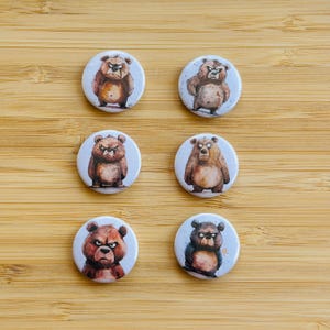 Peut inclure: Six boutons ronds en émail représentant des oursons de dessin animé avec différentes expressions. Les oursons sont bruns et ont des yeux et un nez noirs. Les boutons sont blancs avec une bordure bleu clair.