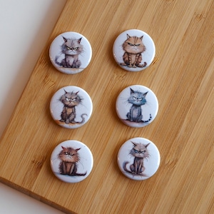 Peut inclure: Six boutons ronds blancs avec des images de chats de dessin animé avec des expressions grincheuses. Les chats sont dans différentes poses et ont des fourrures de différentes couleurs.