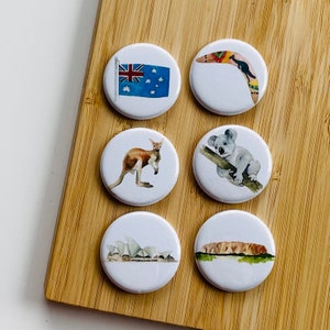 Può includere: Sei bottoni rotondi bianchi con diverse immagini a tema australiano. I bottoni presentano un canguro, un boomerang, un koala, la bandiera australiana, l'Opera House di Sydney e Uluru.