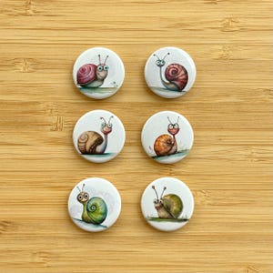 Könnte beinhalten: Sechs runde weiße Knöpfe mit bunten Comic-Schnecken darauf. Jede Schnecke hat eine andersfarbige Schale und lächelt. Die Knöpfe sind in zwei Reihen zu je drei angeordnet.