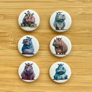 Può includere: Sei bottoni rotondi con diversi ippopotami dei cartoni animati. Gli ippopotami sono tutti di colori diversi e hanno espressioni diverse. I bottoni sono bianchi con un contorno nero.