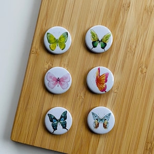 Könnte beinhalten: Sechs runde, weiße, papiergeklebte Magnete mit verschiedenen Schmetterlingsmotiven. Die Schmetterlinge sind grün, rosa, orange und blau.