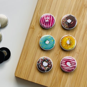 Può includere: Sei magneti colorati a forma di ciambella con diverse guarnizioni, tra cui codette, cioccolato e glassa. I magneti sono disposti in cerchio su una superficie di legno.