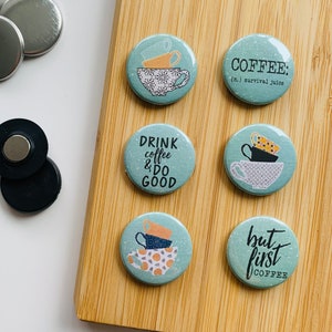 Könnte beinhalten: Sechs runde blaue und weiße Kaffee-Buttons mit verschiedenen Sprüchen. Die Buttons zeigen übereinander gestapelte Kaffeetassen mit weißem Hintergrund und schwarzen und goldenen Akzenten. Die Sprüche auf den Buttons sind "Drink coffee & do good", "Coffee: (n.) survival juice", "But first coffee" und ein Design aus übereinander gestapelten Kaffeetassen.