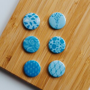 Könnte beinhalten: Sechs runde Anstecknadeln in Blau und Weiß mit verschiedenen Mustern. Die Knöpfe zeigen verschiedene Designs, darunter Anker, Muscheln und Wellen.