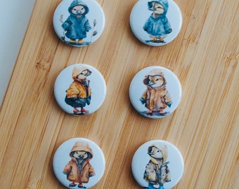 May include: Six round pins with white backgrounds and images of ducks wearing raincoats. The ducks are wearing different colored raincoats, including blue, yellow, and brown.