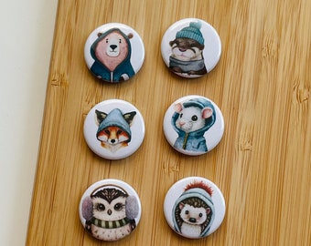 May include: Six round white buttons with colorful animal illustrations. Each button features an animal wearing a hat. The animals include a bear, otter, fox, mouse, owl, and hedgehog.