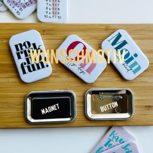Könnte beinhalten: Sortiment an bunten Pins und Magneten mit verschiedenen Sprüchen. Die Pins und Magneten werden auf einer Holzoberfläche präsentiert. Die Sprüche lauten "no risk no fun", "111y day", "Main with kind" und "Kay Sck To".