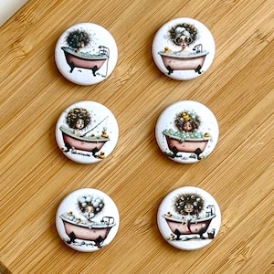 Könnte beinhalten: Sechs weiße runde Buttons mit Illustrationen von Personen in Badewannen. Die Badewannen sind rosa mit schwarzen Klauenfüßen. Die Illustrationen zeigen Personen mit lockigem Haar, Blasen und Gummienten. Die Buttons liegen auf einer Holzoberfläche.