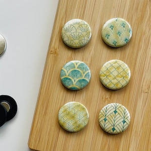 Può includere: Sei magneti rotondi decorativi con una varietà di motivi geometrici in tonalità di giallo e verde. I magneti sono disposti in due file di tre su una superficie di legno.