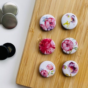 Könnte beinhalten: Sechs runde, weiße Knöpfe mit floralem Muster mit rosa und violetten Blumen. Die Knöpfe sind in zwei Reihen zu je drei auf einer Holzoberfläche angeordnet.