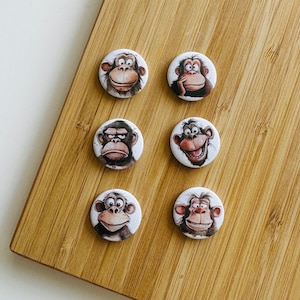 Könnte beinhalten: Sechs runde Knöpfe mit Bildern von Affen darauf. Die Affen haben verschiedene Gesichtsausdrücke. Die Knöpfe sind alle gleich groß und in einem Kreis angeordnet.